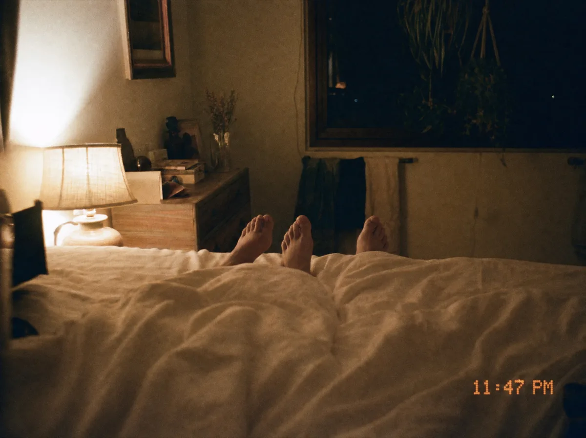 Photograph from the foot of the bed: dim lamplight, feet resting under the covers, completely at ease, timestamped 11:47 PM
