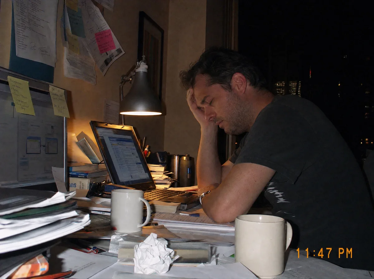 Photograph of an exhausted man at a cluttered desk late at night, head in hand, timestamped 11:47 PM