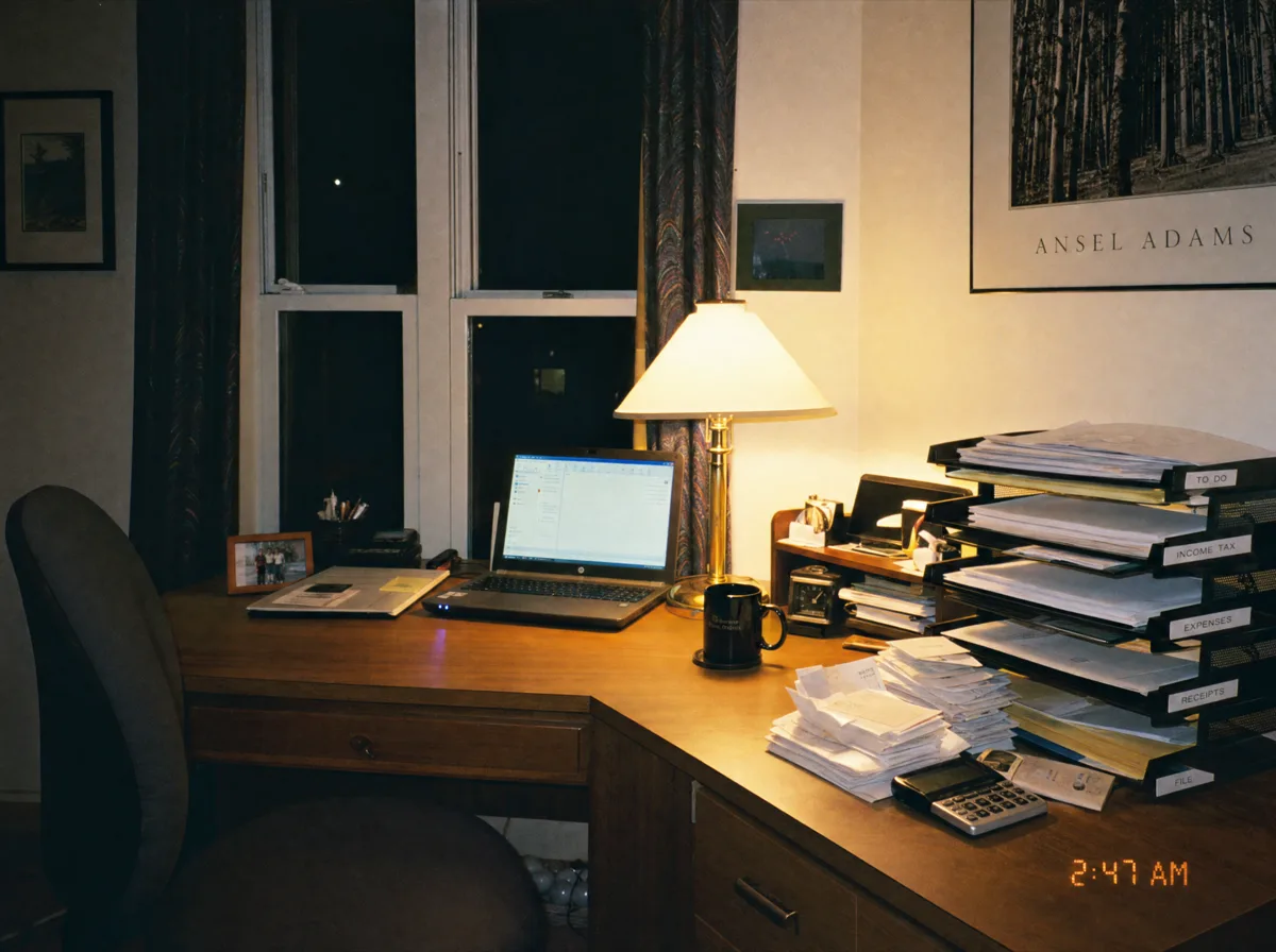 Photograph of a tidy home office desk at night, laptop glowing softly, paperwork filed, nobody in the chair, timestamped 2:47 AM